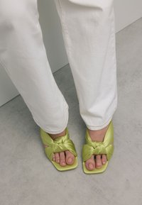 Voeten in limoen groene vierkante teensandalen met gedraaide riemen, gecombineerd met witte broek, staande op een grijze vloer nabij een muur.