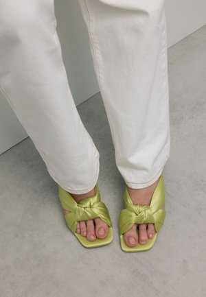 Voeten in limoen groene vierkante teensandalen met gedraaide riemen, gecombineerd met witte broek, staande op een grijze vloer nabij een muur.