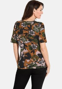 Bluse mit floralem Muster in Braun, Grün und Schwarz, mit lockerer Passform und U-Boot-Ausschnitt. Kombiniert mit schwarzen Hosen.