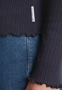 Marineblaues geripptes Stricktop mit Rüschensaum und -manschetten, kombiniert mit blauen Jeans aus Denim mit goldener Stichdetail. Etikett seitlich sichtbar.
