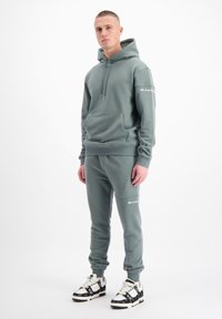 Lichtgroene hoodie en bijpassende joggers van zacht materiaal, met een voorzak en logo-accenten in het wit. Sneakers met zwarte details.