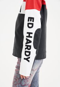 Black and red faux leather jacket featuring a white sleeve with "ED HARDY" text. Its design includes color-blocking and zippered accents.
