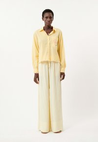 Chemise en lin jaune avec un design de poche, associée à un pantalon beige à jambes larges avec de subtils rayures verticales. Détail d'ourlet effiloché sur la chemise.