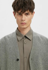 Pelēks kardigans ar pogu aizdari virs brūnas rūtainas krekla. Kardiganam ir mīksta tekstūra un atvērta priekšpuse.