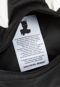 Zwarte ritspouch met een witte binnenvoering en een label. Het label bevat een zwarte silhouet van een figuur en tekst over Jean-Michel Basquiat.