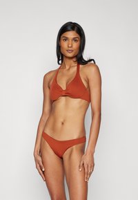 Rūsas krāsas bikini komplekts ar haltera kakla topu, kuram ir mezgla detaļa, un atbilstošiem zema vidukļa apakšdaļām. Gluda auduma ar minimālu šuvju skaitu.