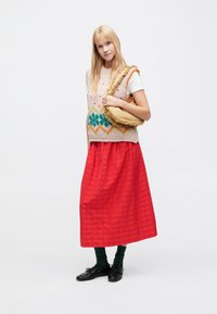 Chaleco de punto con patrones geométricos multicolores, combinado con una falda maxi a rayas rojas. La modelo sostiene un bolso redondo, texturizado y de color marrón claro.