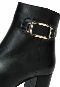 Bottine en cuir noir à bouts carrés, avec un talon épais et une accorte en métal doré sur le côté pour ajouter du détail.