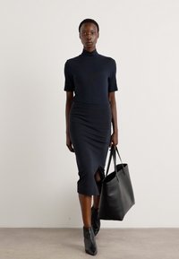 Navy fitted dress with short sleeves and a high neckline, featuring a knee-length skirt with a front slit. Black tote bag and ankle boots.