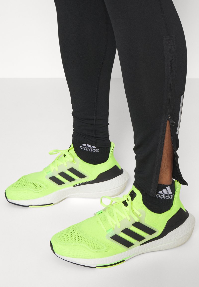 Neon gele sportschoenen met zwarte accenten, voorzien van een ademende bovenkant van mesh en een gevoerde zool. Zwarte leggings met ritssluiting.