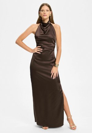 Kobieta ubrana w bezrękawnikową, ciemnobrązową satynową suknię na szyję z rozcięciem do uda i złote sandały na obcasie, pozująca z ręką na biodrze.