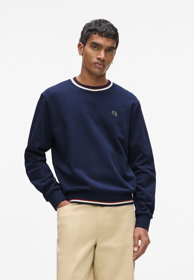 Sweat-shirt bleu marine avec col rond orné de bordures côtes rouges et blanches, manches longues et petit logo brodé sur la poitrine.