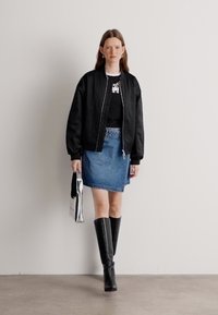 Schwarze Bomberjacke über einem grafischen schwarzen T-Shirt, kombiniert mit einem hellblauen Jeansrock und knielangen schwarzen Stiefeln. Silberne Tasche in der Hand.