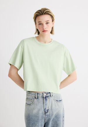 Persoană tânără cu păr blond scurt, purtând un tricou crop verde deschis și blugi albastru șters, cu talie înaltă, stând pe un fundal alb.