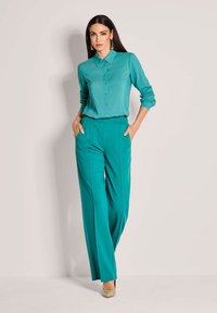 Vrouw staat met handen in de zakken, gekleed in een teal blouse met lange mouwen en bijpassende wijde broek, beige hakken en lange oorbellen.