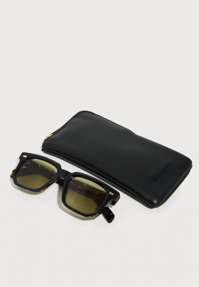 Black square-framed sunglasses with dark lenses beside a black zippered leather case on a white background.