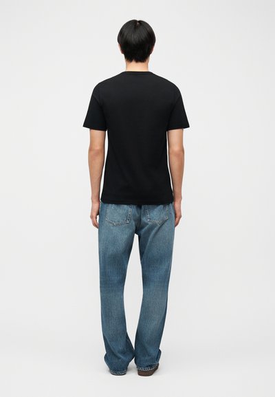 Άτομο που φαίνεται από πίσω, φοράει μαύρο κοντομάνικο μπλουζάκι και άνετο μπλε τζιν, στέκεται μπροστά σε απλό λευκό φόντο.