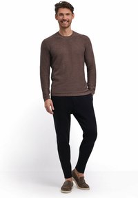 Brauner strukturierter Pullover, schwarze, schmal zulaufende Hosen und graue Slipper. Das Model steht aufrecht vor einem neutralen Hintergrund. Keine weiteren Details sichtbar.