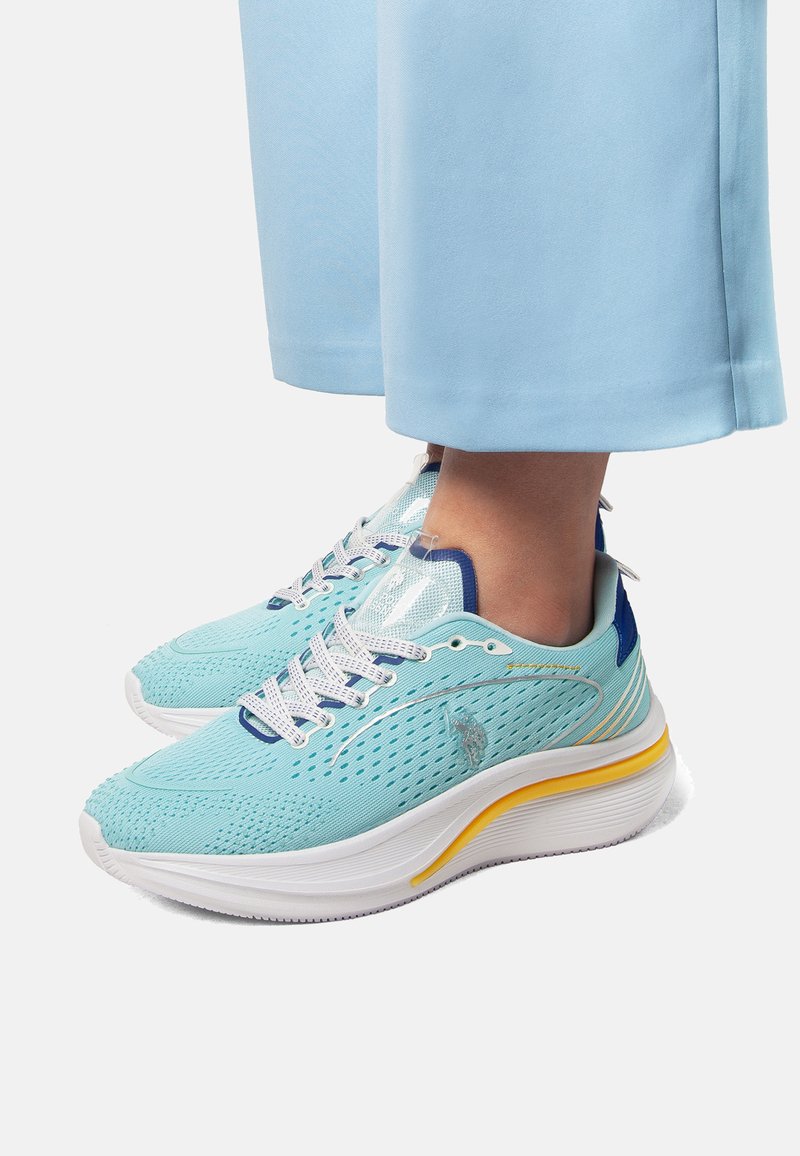 Vaaleansiniset urheilukengät, joissa on teksturoitu verkkomateriaali ylhäällä, valkoinen pohja, keltaiset yksityiskohdat ja sininen kantapää; varustettu nauhoituskiinnityksellä ja logotiedoilla.