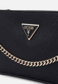Czarna torebka o teksturowanej powierzchni z złotym łańcuchem i trójkątną plakietką Guess USA z znakiem zapytania na środku.