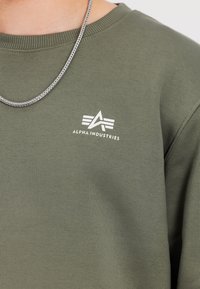 Oliwkowa bluza dresowa z prążkowanym okrągłym dekoltem, z białym logo „ALPHA INDUSTRIES” i fakturowaną tkaniną. Widoczny srebrny łańcuszek.