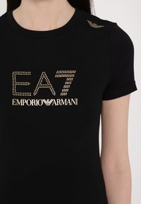 Μαύρη κοντομάνικη μπλούζα με στρογγυλή λαιμόκοψη, featuring χρυσές ανάγλυφες λεπτομέρειες με το λογότυπο "EA7 EMPORIO ARMANI" στο μπροστινό μέρος και διακοσμητικές λεπτομέρειες στους ώμους.