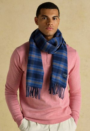 Mężczyzna ubrany w niebiesko-brązowy szalik w kratę, różową koszulę z długim rękawem oraz beżowe spodnie, stojący na jednolitym beżowym tle.