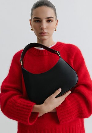 Femeie cu părul pieptănat înapoi, purtând cercei aurii și un pulover roșu, ținând o geantă de mână din piele neagră cu mâner curbat deasupra.