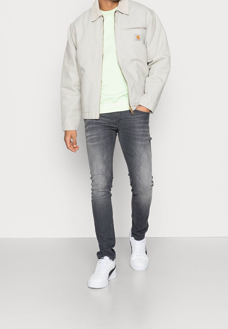Homme portant une veste beige clair, une chemise blanche, un jean gris foncé délavé et des baskets blanches, debout devant un fond uni.