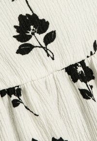 Hvid tekstureret stof med sort blomsterbroderi, der viser synlige blade og kronblade, hvilket skaber et mønster langs en sammensamlet søm.