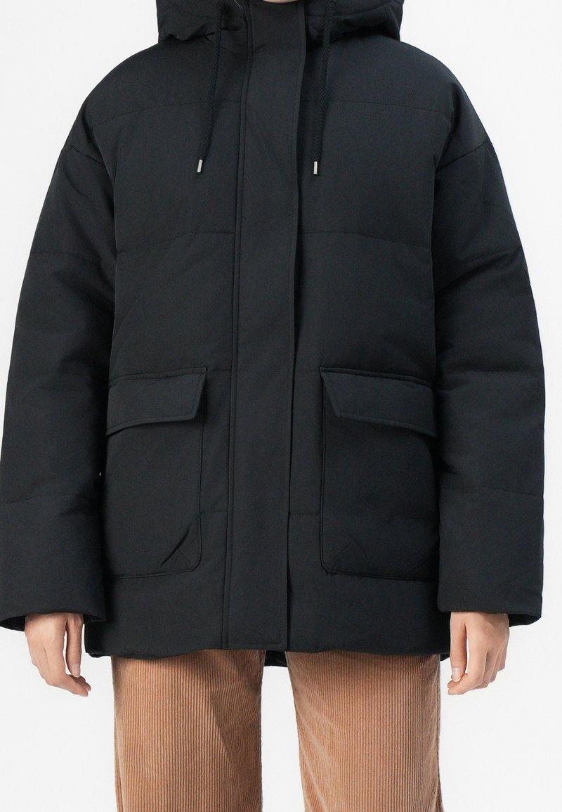 Doudoune noire matelassée avec capuche, dotée d'une fermeture éclair frontale, de deux grandes poches à rabat et d'un motif matelassé. Le matériau semble lisse et mat.