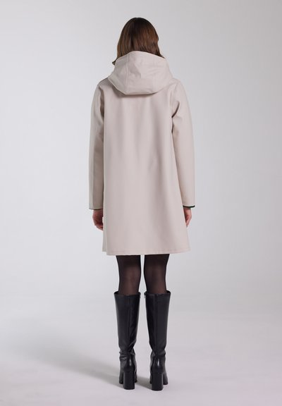 Beige waterdichte jas met een capuchon, halflang, gladde textuur, zijzakken, gecombineerd met zwarte kniehoge haklaarzen en sheer zwarte panty’s.