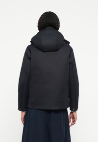 Giacca nera impermeabile con cappuccio, caratterizzata da una texture liscia, polsini elasticizzati e un orlo posteriore leggermente arrotondato. Nessun motivo o accento visibile.