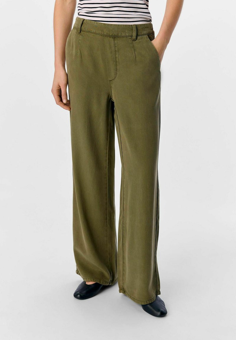 Pantalones de pierna ancha en tela verde oliva con una textura suave, cuentan con una cinturilla y bolsillos laterales. Combinados con calzado negro.