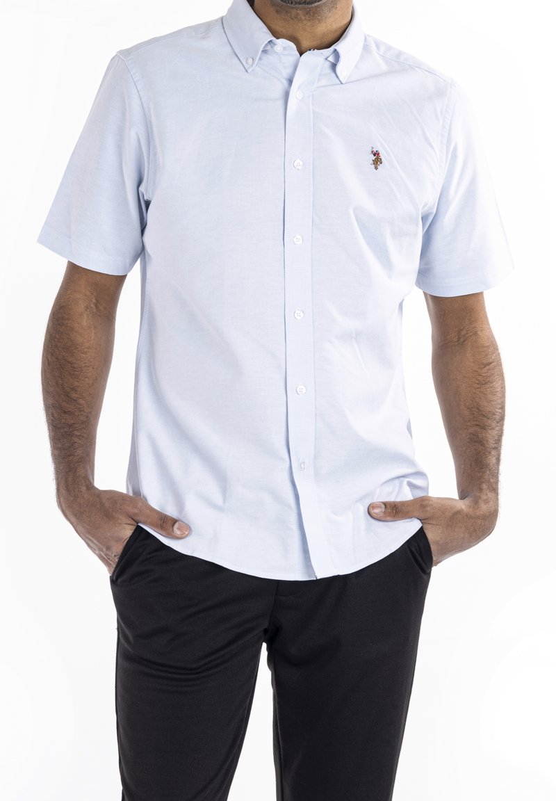 Camisa de manga corta de color azul claro con botones y cuello, con un logo bordado en el pecho izquierdo, combinada con pantalones negros. Textura suave.