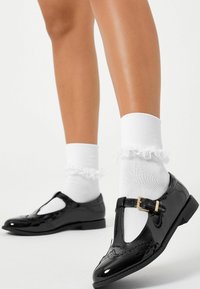 Svarta lackskinn Mary Jane-skor med T-rem, gyllene spänne och intrikata detaljer, kombinerat med vita volangstrumpor som går upp till ankeln.