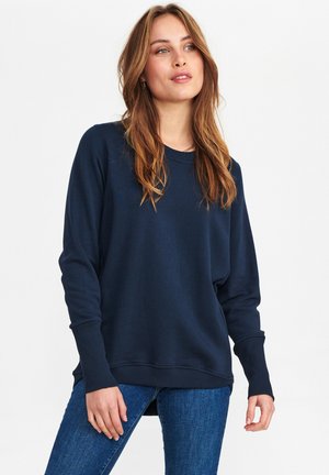 Granatowy sweter z miękkiego materiału, luźny krój, ściągacze przy mankietach, okrągły dekolt, boczne rozcięcia. W połączeniu z niebieskimi dżinsami, wyeksponowany na białym tle.