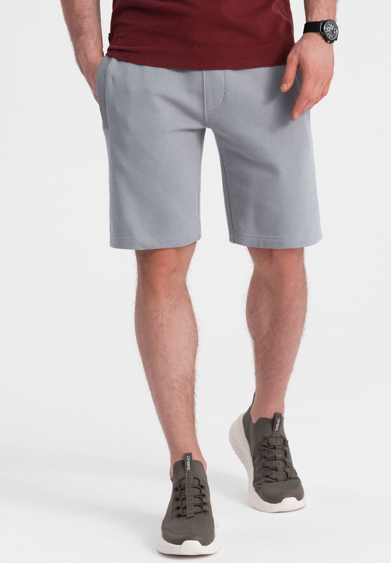 Pantalones cortos de sudadera grises con un corte relajado, que cuentan con bolsillos laterales y una textura suave, combinados con deportivas verde oliva con suelas blancas.
