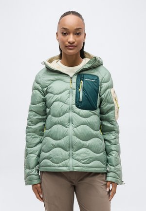 Žena oblečená v svetlozelenej prešívanej bunde s tmavozelenou náprsnou kapsou a béžovými akcentmi na zips, stojaca pred jednoduchým pozadím.