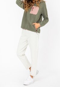 Chaqueta de forro polar en verde oliva con un bolsillo en el pecho rosa y cremallera. Combinada con pantalones blancos y zapatillas blancas, con puños remangados.