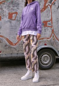 Fioletowa bluza z kapturem z przednią kieszenią i białym logo, zestawiona z wzorzystymi spodniami z wirami w kolorach beżu, żółci i szarości. Białe wysokie trampki.