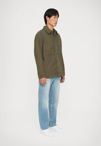 Camicia di velluto a coste verde oliva con due tasche frontali, chiusura con bottoni e colletto, abbinata a jeans dritti blu chiaro e sneakers bianche.