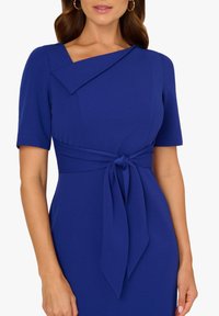 Niebieska sukienka z gładkiego materiału, z krótkimi rękawami, asymetrycznym dekoltem i wiązaną talią, podkreślającą sylwetkę.