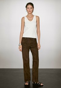 Vrouw in een witte mouwloze top, bruine suède broek, zwarte sandalen en zilveren armbanden, staande tegen een neutrale achtergrond.