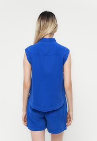 Blaues ärmelloses Hemd mit Kragen, das einen abgerundeten Saum und Stickdetails aufweist. Kombiniert mit passenden blauen Shorts.