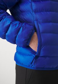 Main dans la poche latérale d'une veste matelassée bleu vif portée avec un bas noir, visible de la taille jusqu'au milieu du bras.