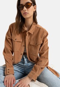 Bruine corduroy shirt jacket met twee voorzakken, knoopsluitingen en lange mouwen. G gedragen over lichtblauwe spijkerbroek. Accessoires omvatten een paar bruine zonnebrillen.