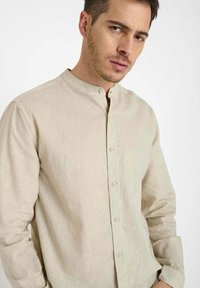 Jonge man in een beige langslavige overhemd met Mandarin-kraag, kijkend naar de camera met een neutrale uitdrukking.