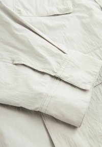 Chaqueta color beige claro con tejido texturizado, mangas dobladas y detalles de costura visibles. Diseño simple y minimalista sin adornos.