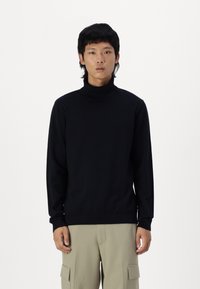 Jersey de cuello alto negro con mangas largas y puños acanalados. Hecho de una tela suave, diseñado para ajustarse cómodamente, usado con pantalones de color claro.